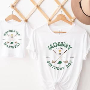 May include: Two white t-shirts with green text and graphics. The larger shirt says "Mommy of the Birthday Boy" with a golf club and a trophy. The smaller shirt says "Birthday Boy" with a golf club and a golf hat. The text on both shirts says "Est. 2023".
