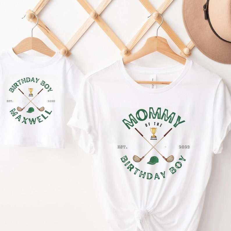 May include: Two white t-shirts with green text and graphics. The larger shirt says "Mommy of the Birthday Boy" with a golf club and a trophy. The smaller shirt says "Birthday Boy" with a golf club and a golf hat. The text on both shirts says "Est. 2023".