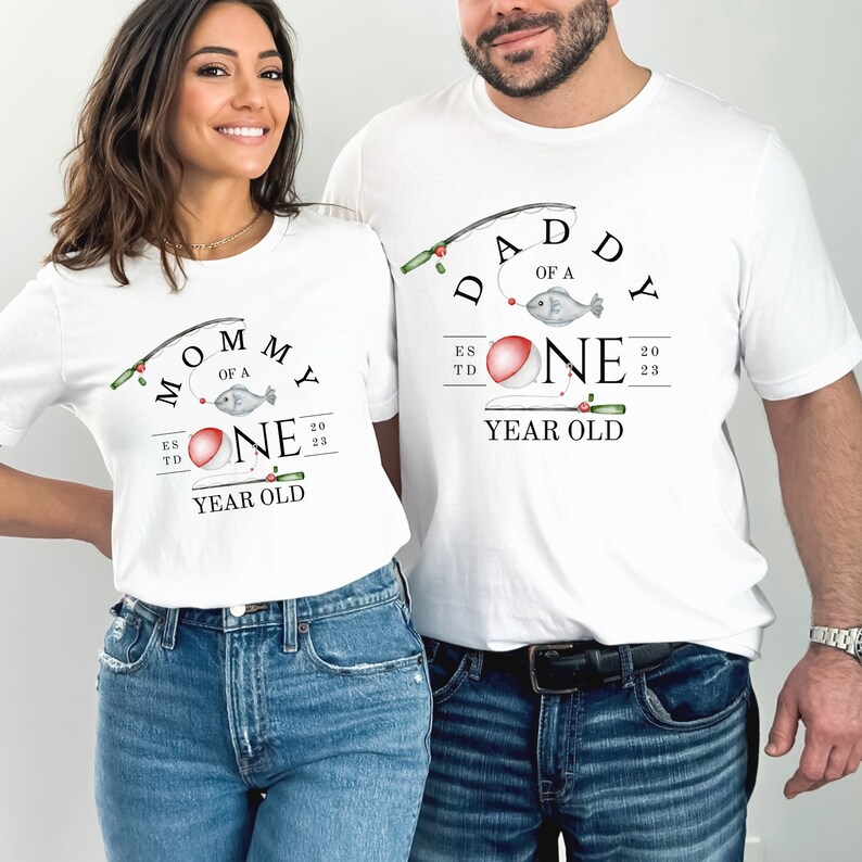 May include: Two white t-shirts with a fishing theme. The shirt on the left says "Mommy of a One Year Old" with a red bobber and a fish on a fishing line. The shirt on the right says "Daddy of a One Year Old" with a red bobber and a fish on a fishing line.