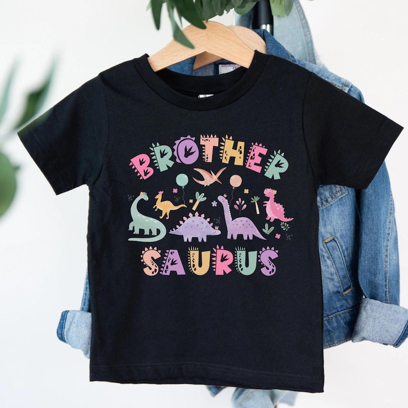 K&ouml;nnte beinhalten: Schwarzes T-Shirt mit dem Text "Brother Saurus" in bunten Buchstaben und Dinosaurier-Illustrationen. Das Design umfasst verschiedene Dinosaurier und dekorative Elemente, perfekt f&uuml;r ein junges Kind.