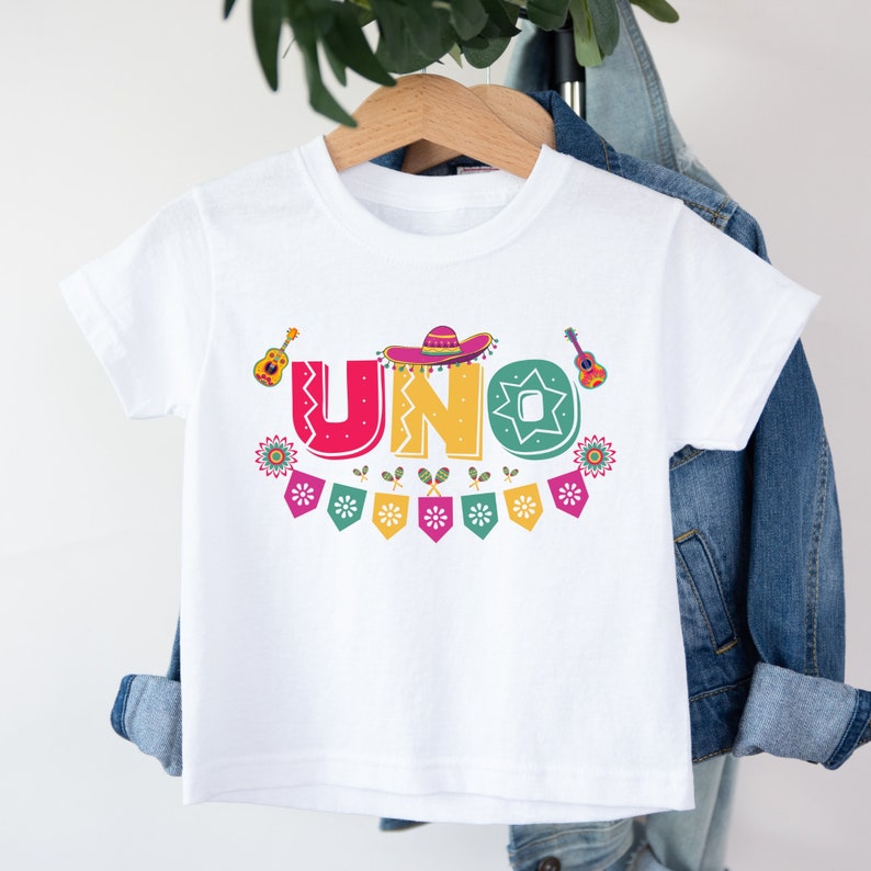 K&ouml;nnte beinhalten: Wei&szlig;es T-Shirt mit einer bunten Grafik des Wortes "UNO" im festlichen Stil. Die Grafik enth&auml;lt einen Sombrero, Maracas und Gitarren.