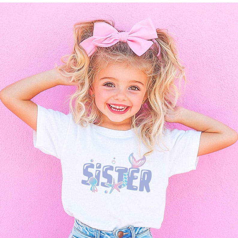 May include: A young girl with blonde hair wearing a white t-shirt with the word "Sister" in blue and purple letters with a mermaid tail, starfish, and jellyfish graphic. She is wearing a pink bow in her hair.