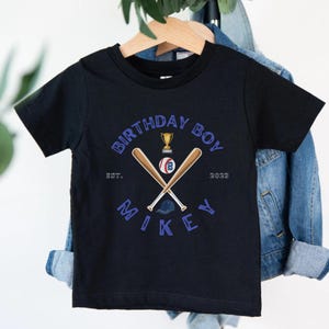 K&ouml;nnte beinhalten: Schwarzes kurz&auml;rmeliges T-Shirt mit Baseball-Design. Das Design umfasst gekreuzte Baseballschl&auml;ger, einen Baseball, eine Troph&auml;e und den Text "BIRTHDAY BOY" und "MIKEY". Das Jahr "2023" ist ebenfalls enthalten.