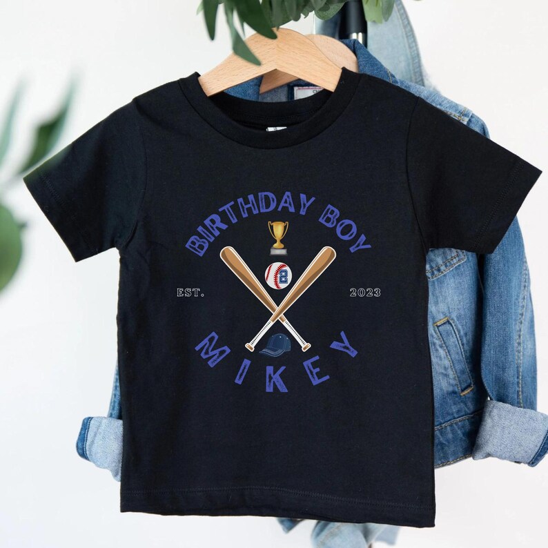 K&ouml;nnte beinhalten: Schwarzes kurz&auml;rmeliges T-Shirt mit Baseball-Design. Das Design umfasst gekreuzte Baseballschl&auml;ger, einen Baseball, eine Troph&auml;e und den Text "BIRTHDAY BOY" und "MIKEY". Das Jahr "2023" ist ebenfalls enthalten.