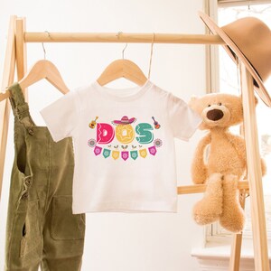 Peut inclure: T-shirt blanc pour enfant avec un graphisme color&eacute; du mot "DOS" avec un sombrero, une guitare et des drapeaux festifs.