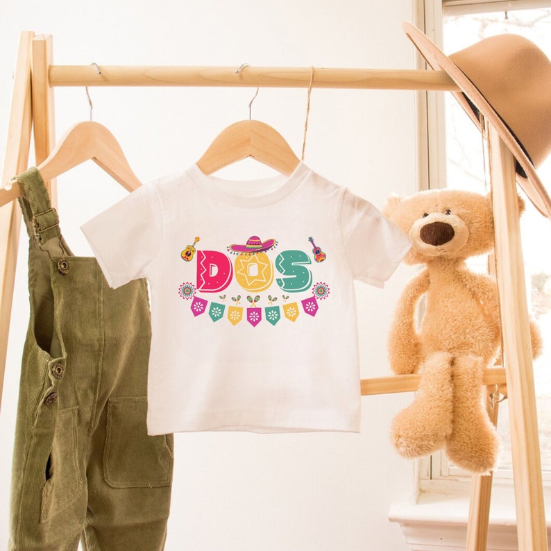 Peut inclure: T-shirt blanc pour enfant avec un graphisme color&eacute; du mot "DOS" avec un sombrero, une guitare et des drapeaux festifs.