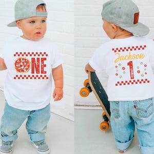 May include: White t-shirt featuring a pizza graphic and the word "ONE" in red, with a checkered border. The back of the shirt displays the name "Jackson" and the number "1" with pizza slice graphics. The child wears blue jeans and a grey baseball cap.