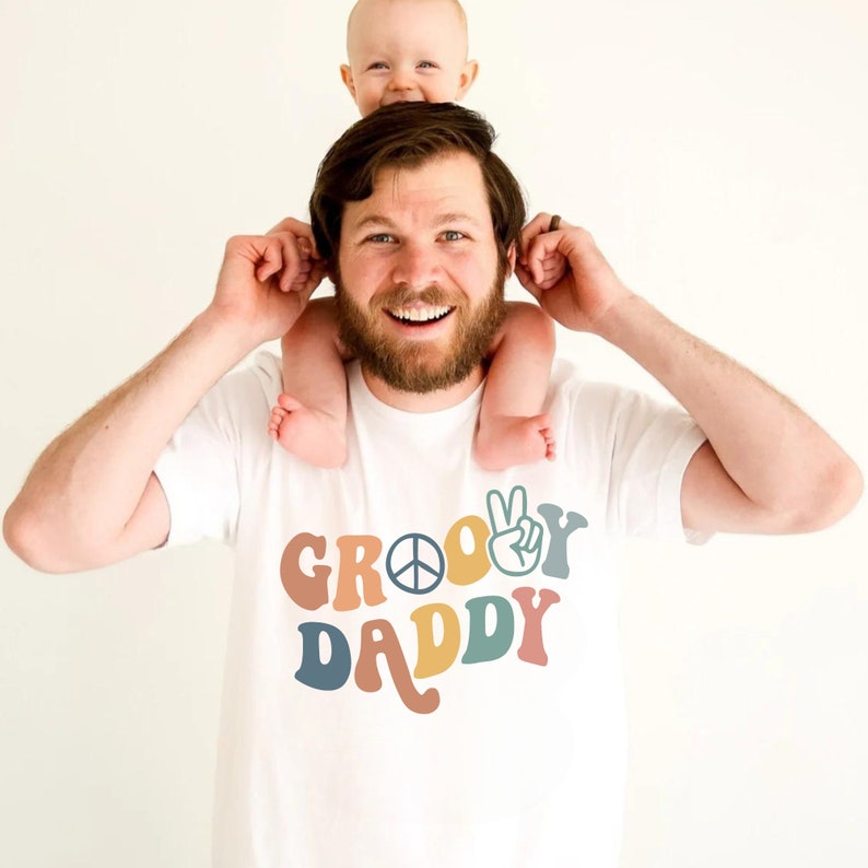 May include: A white t-shirt with the text "Groovy Daddy" in colorful retro font. A peace sign and a peace hand gesture are included in the design. A baby is sitting on the shoulders of a person wearing the shirt.