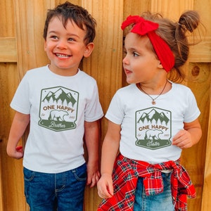 May include: Two children wearing white t-shirts with a green mountain and bear graphic. The boy's shirt says "One Happy Brother Est. 2022" and the girl's shirt says "One Happy Sister Est. 2022".