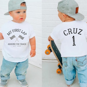 Peut inclure: Un t-shirt blanc avec le texte "MY FIRST LAP" et des drapeaux à damier croisés. Le t-shirt dit aussi "AROUND THE TRACK - EST 2024". L'enfant porte un jean bleu clair et une casquette de baseball grise. Le dos du t-shirt dit "CRUZ 1".