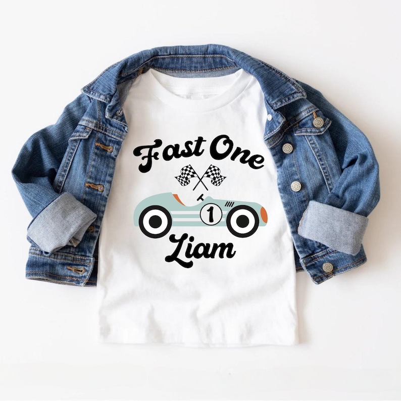 May include: White t-shirt with a blue race car graphic and the text "Fast One" above the car and "Liam" below the car. The car has a number "1" on the side and checkered flags above it.