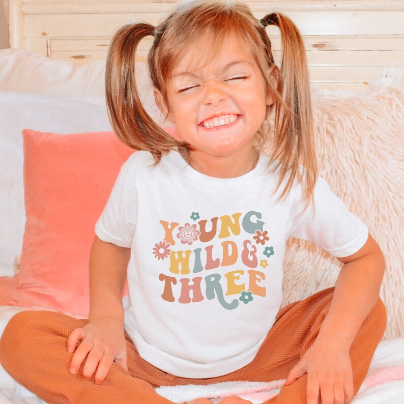 May include: A white t-shirt with a colorful graphic that says "Young Wild & Three" with flowers and a peace sign. The shirt is being worn by a young child with blonde hair.