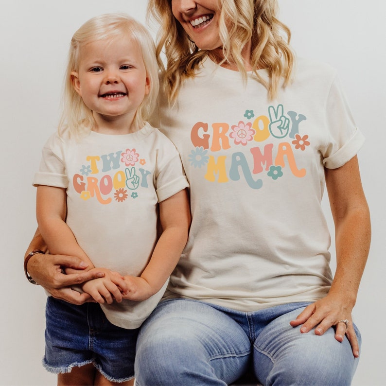 May include: Two matching t-shirts, one with the text "Two Groovy" and a peace sign, and the other with the text "Groovy Mama" and a peace sign. Both shirts are cream colored with colorful retro-style lettering and flowers.