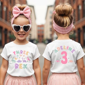 May include: A white t-shirt featuring the words "THREE REX" in pastel colours and dinosaur illustrations. The back of the shirt displays the name "ADELINE" and the number "3". The girl wears a pink skirt, heart-shaped sunglasses, and a pink gingham bow.