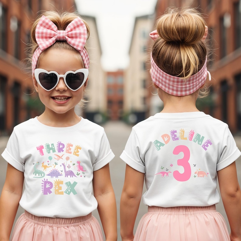 May include: A white t-shirt featuring the words "THREE REX" in pastel colours and dinosaur illustrations. The back of the shirt displays the name "ADELINE" and the number "3". The girl wears a pink skirt, heart-shaped sunglasses, and a pink gingham bow.