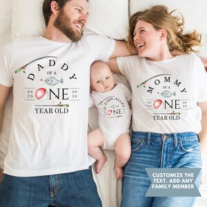 May include: A white t-shirt with the text "Daddy of a One Year Old" and a fishing rod with a red bobber and a fish. The text "Est. 2023" is also on the shirt. A white onesie with the text "O-Fish-Alley One" and a fishing rod with a red bobber and a fish. The text "Est. 2023" is also on the onesie. A white t-shirt with the text "Mommy of a One Year Old" and a fishing rod with a red bobber and a fish. The text "Est. 2023" is also on the shirt.