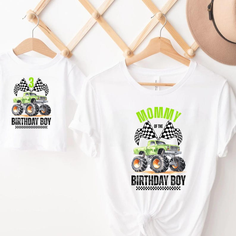 Puede incluir: Camisetas blancas con un dise&ntilde;o de monster truck. Una camiseta dice "3 BIRTHDAY BOY" con un monster truck verde y banderas a cuadros. La otra dice "MOMMY OF THE BIRTHDAY BOY". Ambas est&aacute;n en perchas de madera.