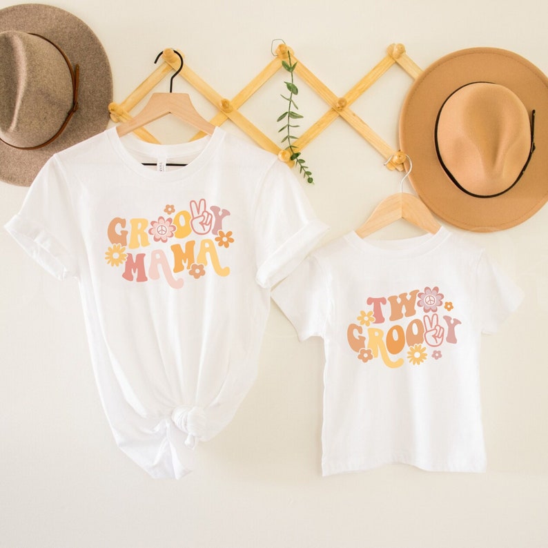 May include: Two white t-shirts with a groovy theme. The larger shirt says "Groovy Mama" with peace signs and flowers. The smaller shirt says "Two Groovy" with peace signs and flowers.