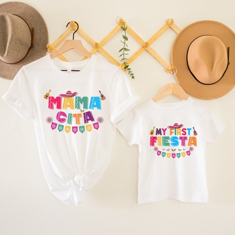 K&ouml;nnte beinhalten: Zwei wei&szlig;e T-Shirts mit buntem Text und Grafiken. Das gr&ouml;&szlig;ere Shirt sagt "Mama Cita" mit einem Sombrero, einer Gitarre und festlichen Flaggen. Das kleinere Shirt sagt "My First Fiesta" mit einem Sombrero, einer Gitarre und festlichen Flaggen.