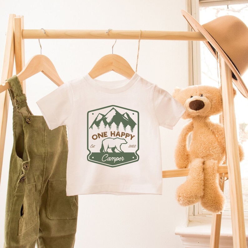 May include: White baby t-shirt with a green and brown graphic of a bear in front of mountains. The graphic says "One Happy Camper" with the year 2022 and "Est" above the bear.
