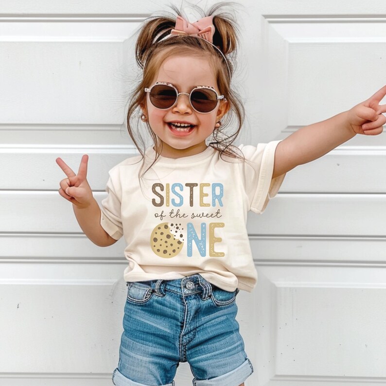 May include: A young child wearing a cream-colored t-shirt with the words "SISTER of the sweet ONE" and a cookie graphic. The child is also wearing denim shorts and sunglasses, and is smiling while making a peace sign.