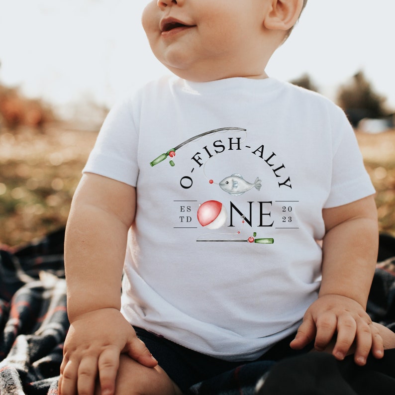 May include: A white baby onesie with a fishing-themed graphic that says "O-Fish-Ally One" with a red and white bobber, a fish, and fishing rods.