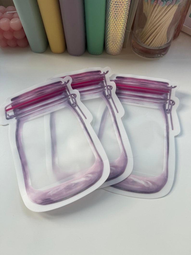 May include: Two clear plastic reusable storage bags with a purple and pink mason jar design. The bags have a zipper closure and are ideal for storing food or other small items.