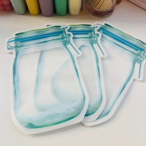 May include: Three clear plastic reusable food storage bags with a blue zipper and a printed design of a glass jar with a lid.