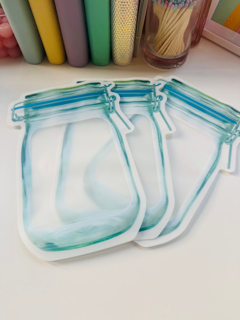 May include: Three clear plastic reusable food storage bags with a blue zipper and a printed design of a glass jar with a lid.