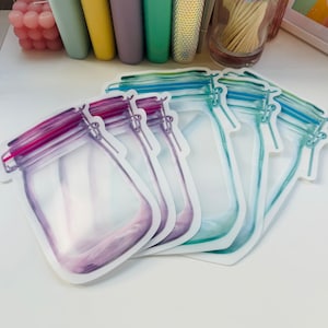 May include: Five reusable food storage bags in the shape of mason jars. The bags are clear with a white border and have a zipper closure. The bags are in different colors, including purple, blue, and green.