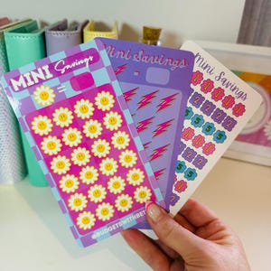 May include: Three colourful sticker sheets with the text "Mini Savings" on each sheet. The first sheet has a pink background with white daisies and yellow smiley faces. The second sheet has a purple background with pink lightning bolts. The third sheet has a purple background with pink and blue circles with numbers.