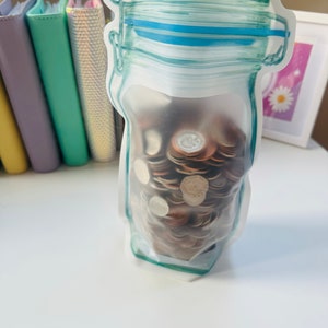 May include: A clear plastic bag shaped like a glass jar filled with a stack of coins. The bag has a blue and green design.
