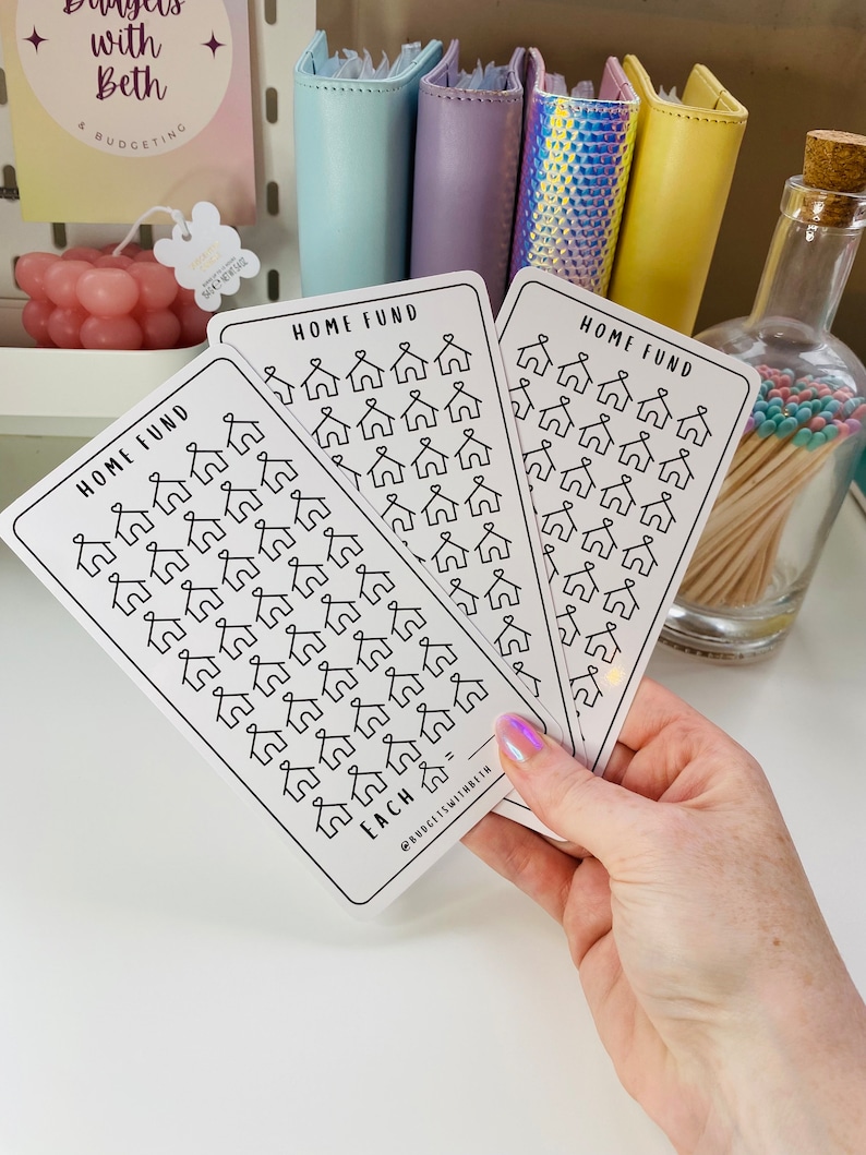 May include: Three white cards with black outlines of houses printed on them. Each card has the text "HOME FUND" at the top and the text "EACH" at the bottom. The cards are held in a hand.