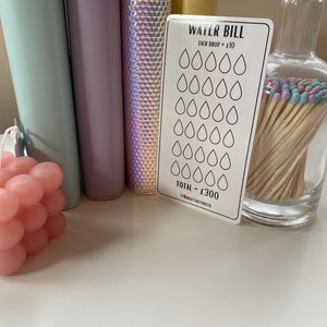 May include: A white card with the text "WATER BILL" and a grid of 30 water drops. Each drop is worth $10. The total bill is $300. The card is on a white surface with a pink candle, a glass jar with matches, and four colorful binders.