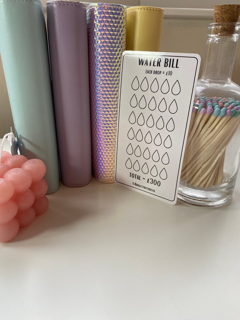 May include: A white card with the text "WATER BILL" and a grid of 30 water drops. Each drop is worth $10. The total bill is $300. The card is on a white surface with a pink candle, a glass jar with matches, and four colorful binders.