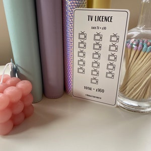 May include: A white sticker with a black border and text that reads "TV LICENCE". The sticker has a grid of 16 television icons with a checkmark in each icon. The text "EACH TV = &pound;10" and "TOTAL - &pound;160" is printed on the sticker. The sticker is on a white surface.