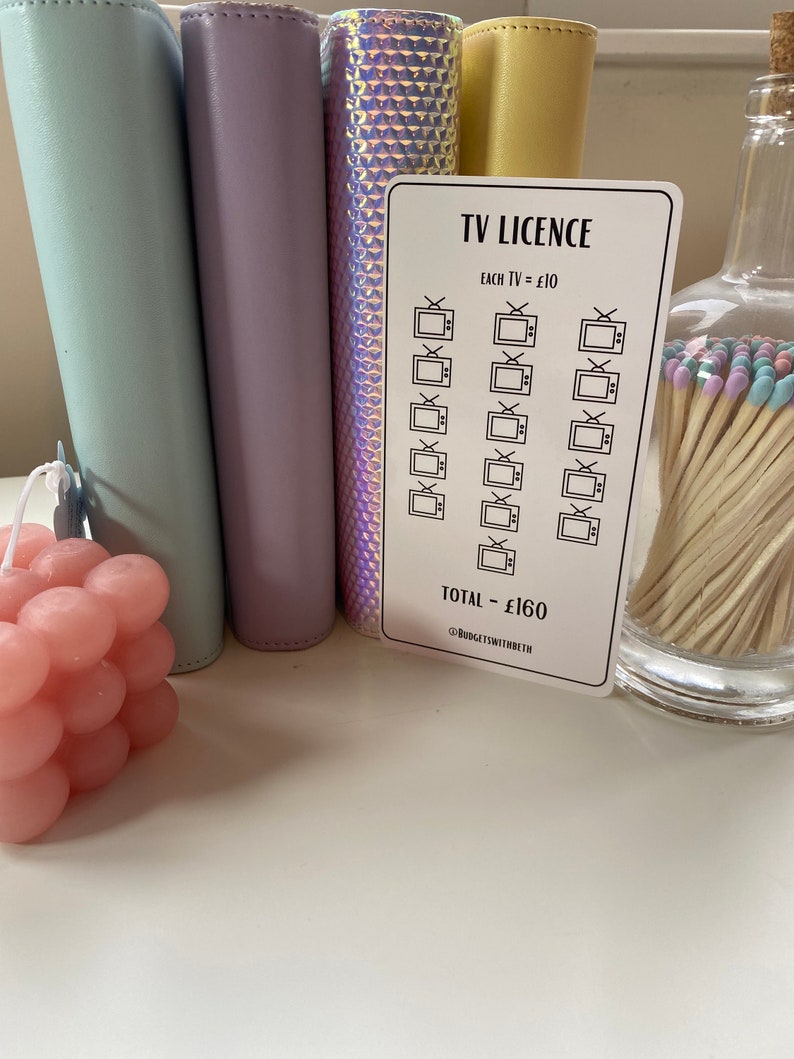 May include: A white sticker with a black border and text that reads "TV LICENCE". The sticker has a grid of 16 television icons with a checkmark in each icon. The text "EACH TV = &pound;10" and "TOTAL - &pound;160" is printed on the sticker. The sticker is on a white surface.
