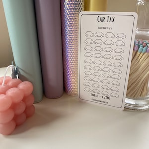 May include: A white card with the text "CAR TAX" and a grid of 40 small car icons. Each car icon is worth &pound;5 and the total is &pound;200. The card is on a table with a pink candle, a jar of matches, and four colorful binders.