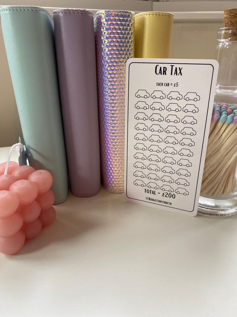 May include: A white card with the text "CAR TAX" and a grid of 40 small car icons. Each car icon is worth &pound;5 and the total is &pound;200. The card is on a table with a pink candle, a jar of matches, and four colorful binders.