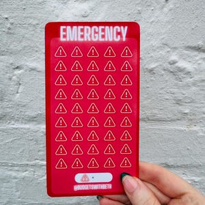 May include: A red rectangular card with the word "EMERGENCY" in white, bold letters at the top. The card features numerous white exclamation point symbols inside red triangles. The bottom of the card has the text "@BUDGETSWITHBETH".