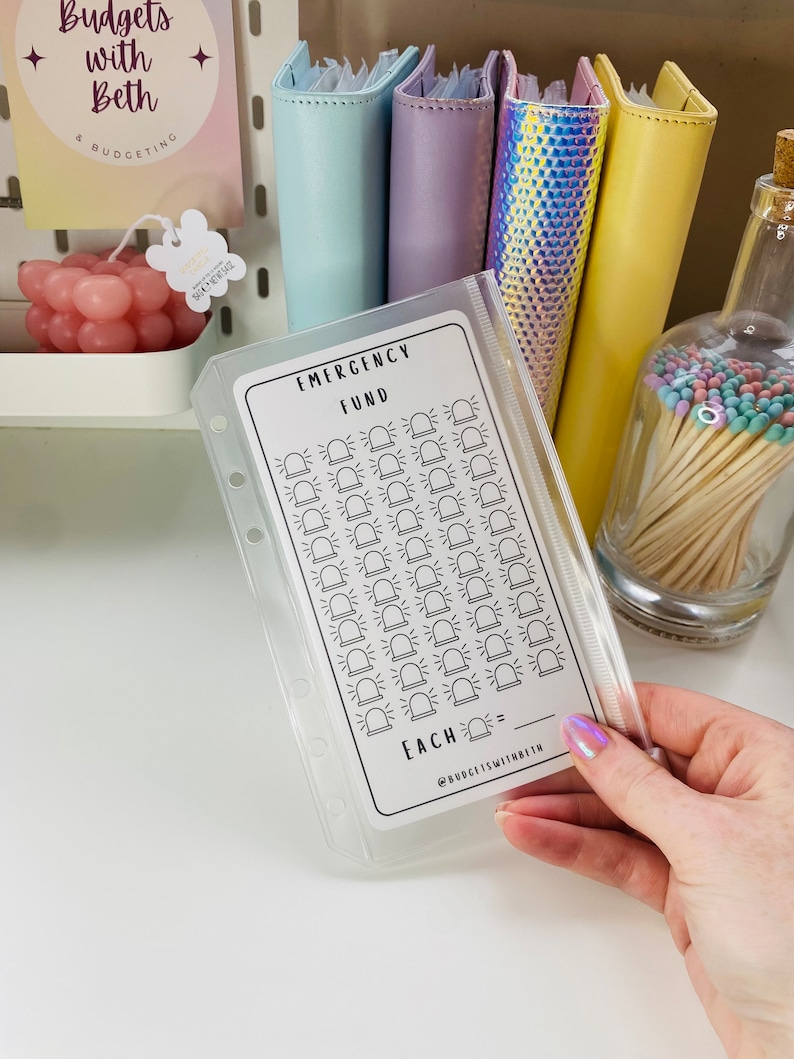 May include: A white printable emergency fund tracker with a black outline of a siren. The tracker has 50 spaces for each dollar saved. The text "EMERGENCY FUND" and "EACH =" are printed on the tracker.