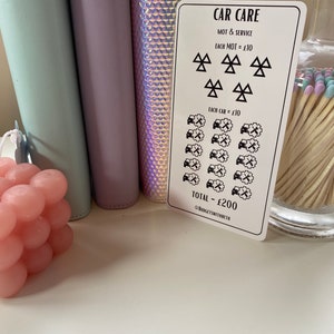 May include: A white card with black text and symbols outlining the cost of car care. The card reads "CAR CARE", "MOT & SERVICE", "EACH MOT = &pound;10", "EACH CAR = &pound;10", and "TOTAL - &pound;200". The card also includes a symbol for a car with a wrench and a symbol for an MOT test.