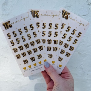 May include: Three white cards with gold text and numbers are held. The cards say "1K savings!" and feature gold numbers 5, 10, and 20. Gold stars and hearts are also printed on the cards. The cards are against a white brick wall.