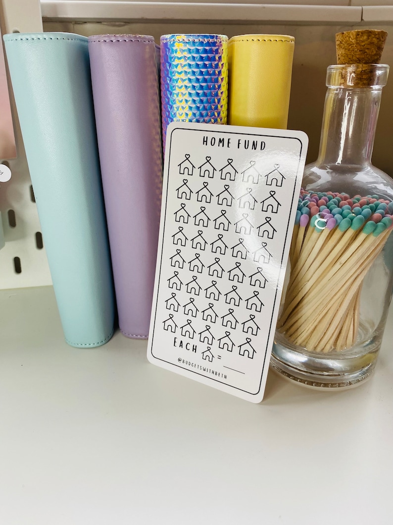 May include: A white card with black text that says "Home Fund" and has a grid of 36 small house icons. The text below the grid says "Each =" with a blank line for writing. The text at the bottom of the card says "@budget.is.within.reach".