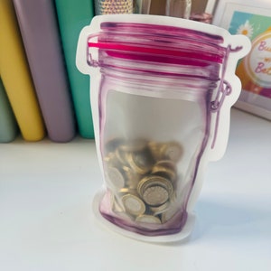May include: A clear plastic reusable food storage bag shaped like a glass jar with a pink lid. The bag is filled with gold-colored coins.