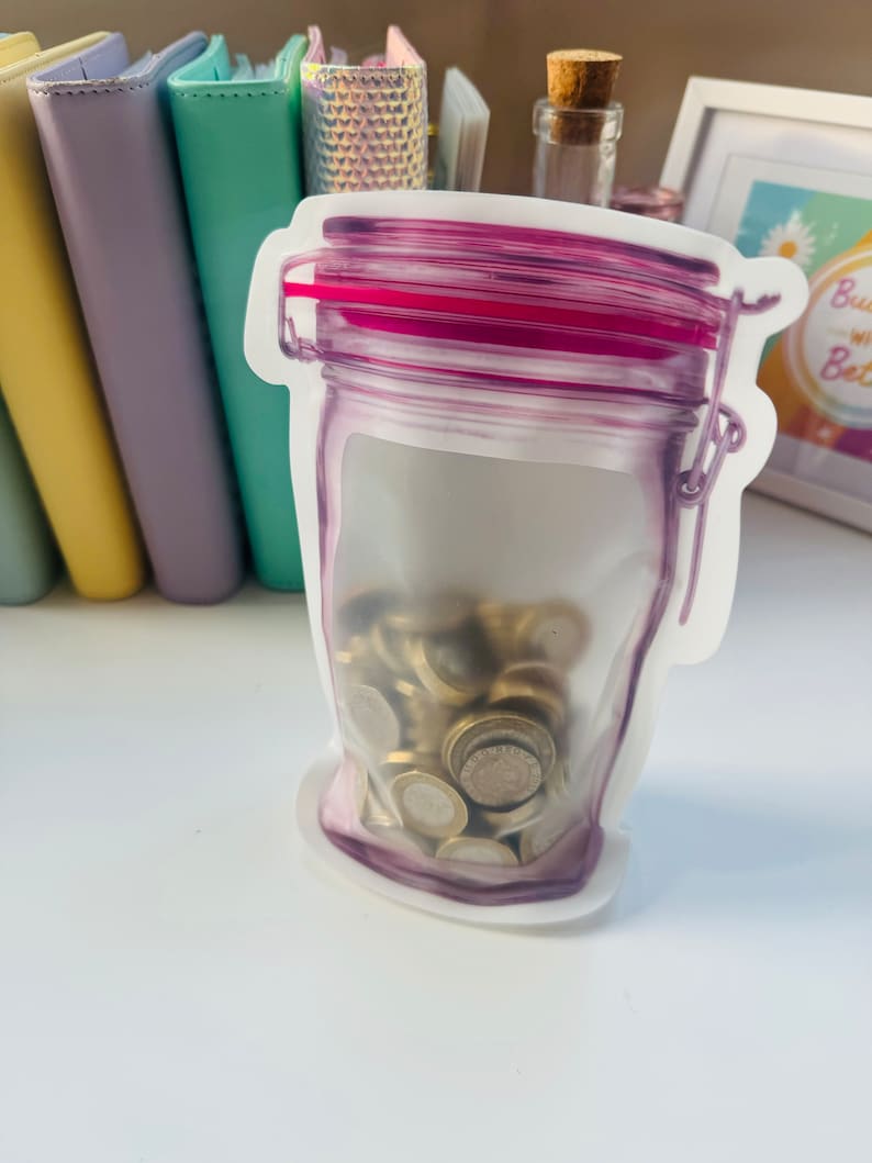 May include: A clear plastic reusable food storage bag shaped like a glass jar with a pink lid. The bag is filled with gold-colored coins.