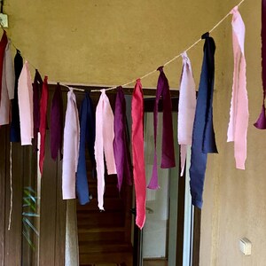 Op de afbeelding: Een kleurrijke stoffen slinger met verschillende tinten roze, rood, paars en marineblauw. De stoffen stroken zijn aan een touw opgehangen en vormen een decoratieve banner. De slinger hangt voor een houten deur.