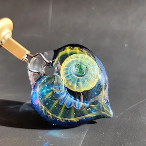 Op de afbeelding: Een glazen pijp met een blauw, groen en geel swirl-ontwerp. De pijp heeft een ronde, heldere kamer met een klein gaatje aan de zijkant. De pijp is bevestigd aan een gouden metalen steel.