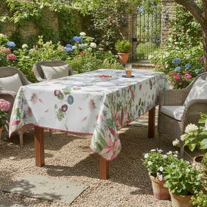 Könnte beinhalten: Ein rechteckiger Holztisch, bedeckt mit einer weißen Blumendecke. Die Tischdecke hat ein Muster aus rosa, grünen und lila Blumen. Zwei Korbstühle mit cremefarbenen Kissen stehen auf beiden Seiten des Tisches. Die Szene spielt in einem Garten.