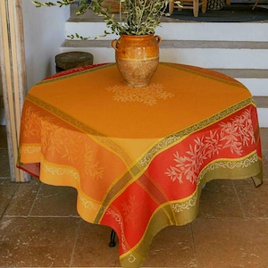 Könnte beinhalten: Eine quadratische orangefarbene Tischdecke mit floralem Design und rotem Rand. Die Tischdecke ist über einem runden Tisch drapiert. Eine Keramikvase mit einer Pflanze steht in der Mitte des Tisches.